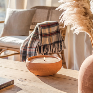 Handgemaakte terracotta schaal gevuld met kaarsenzand en een brandende lont, geplaatst op een houten tafel in een warme, bohemien interieursetting met een plaid en pampasgras.