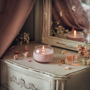 Een sfeervolle setting met een brandende roze kaars in een glazen kom op een vintage witgelakte kaptafel, omringd door rozenblaadjes en een klein parfumflesje. Op de achtergrond is een spiegel zichtbaar waarin de kaars en bloemendecoratie weerspiegeld worden.