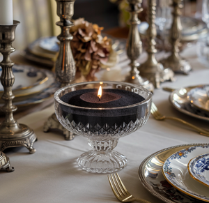 Elegant gedekte tafel met een brandende kaars in zwart kaarsenzand, gepresenteerd in een kristallen coupeglas. De tafel is versierd met zilveren kandelaars, porseleinen borden met bloemenmotief en gouden bestek, wat zorgt voor een luxueuze en feestelijke sfeer.
